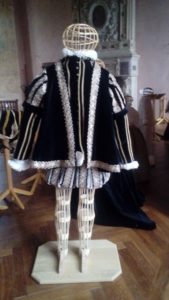 Costume historique reconstitué masculin vers 1550-1560 composé d'un manteau noir à manches courtes à crevés orné de passementerie dorée, d'un pourpoint noir orné de passementerie dorée, d'un haut-de-chausses gris clair avec des passementeries noires et dorées et d'une chemise à ruchés au col et aux poignets