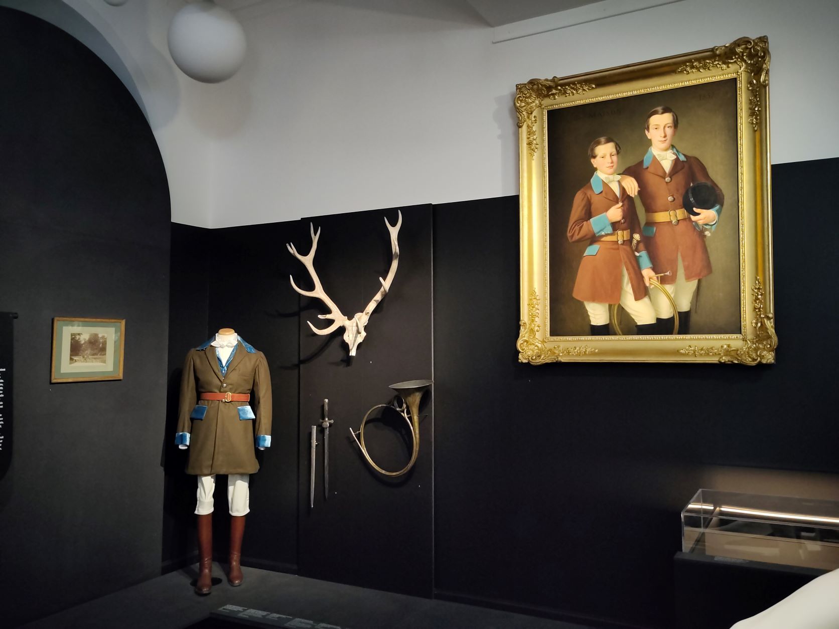 Vue de l'exposition "Châteaux de l'Aube". La tenue de chasse est présentée avec une dague, un crâne de cerf, un cor de chasse et le portrait de Fernand et Angelo de Taisne.
