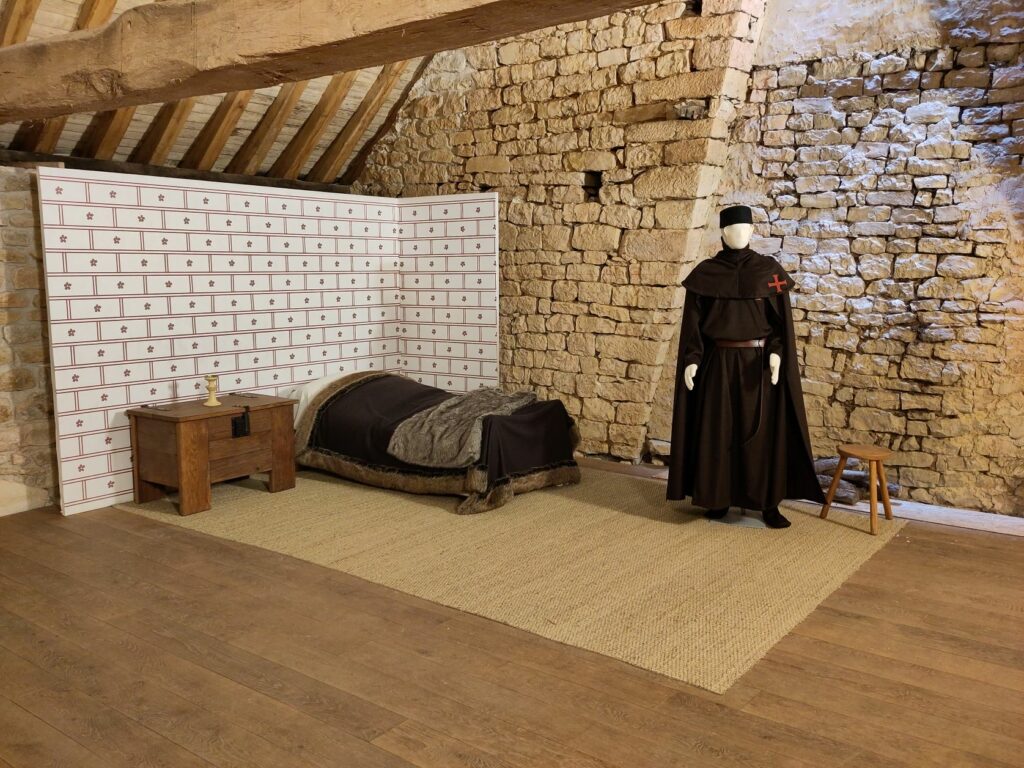 Chambre reconstituée avec un lit, un coffre, un mur peint, un tapis et un mannequin portant une tenue de templier noir