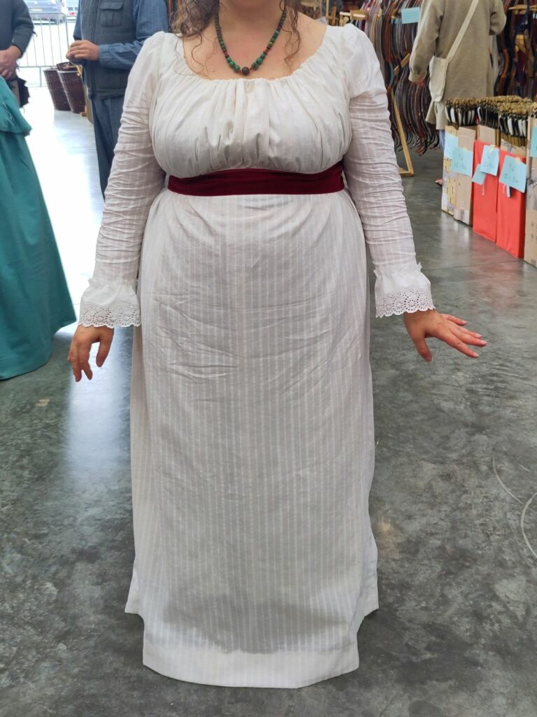 Robe blanche ceinturée sous la poitrine par un large ruban de velours rouge, les bas de manches sont en broderie anglaise froncée