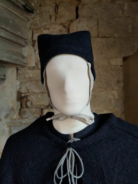 Tête du mannequin habillé d'un bonnet noué sous le menton et d'un petit chapeau gris foncé. Vue de face