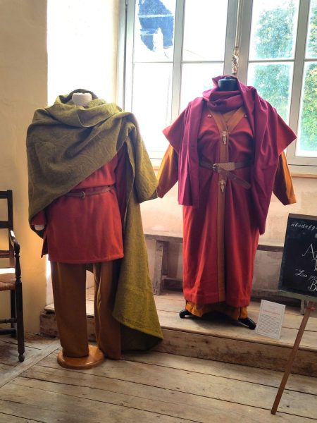Deux costumes portés sur des bustes d'exposition. Une tenue masculine dans des tons orangés avec un manteau drapé vert. Une tenue féminine dans des tons rouges et orangés.