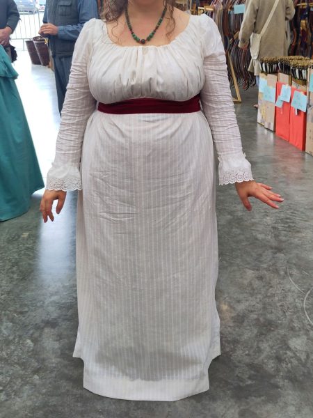Robe blanche ceinturée sous la poitrine par un large ruban de velours rouge, les bas de manches sont en broderie anglaise froncée