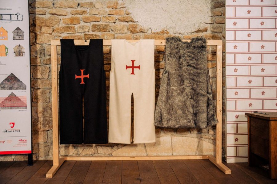 Un portant en bois sur lequel sont supensdus par les épaules : un tabard marron à croix rouge, un tabard blanc à croix rouge et un pelisson en fausse fourrure grise
