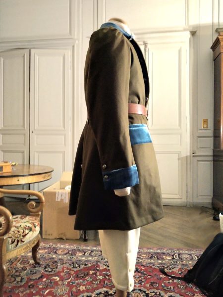 Reconstitution de la tenue de chasse vue de profil avec la redingote en lainage marron aux détails en velours turquoise, la culotte blanche et une ceinture en cuir